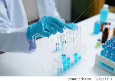 Scientist Conducting Experiment with Test Tubes in Laboratory Wearing Blue Gloves Scientist Conducting Experiment with Test Tubes in Laboratory Wearing Blue Gloves 128861344