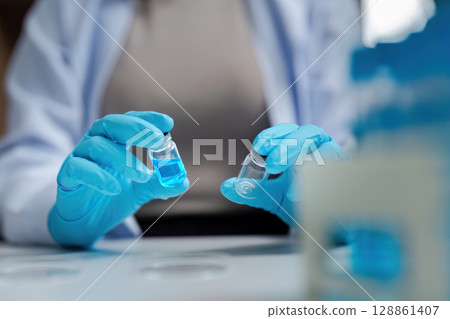 Scientist in Laboratory Holding Vials with Blue Liquid, Wearing Protective Gloves and Lab Coat, Conducting Research 128861407
