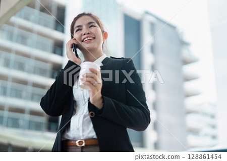 Businesswoman Smiling While Talking on Phone and Holding Coffee Cup in Modern Urban Setting Businesswoman Smiling While Talking on Phone and Holding Coffee Cup in Modern Urban Setting 128861454