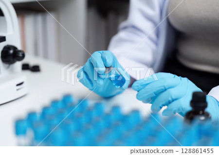 Scientist in Laboratory Handling Blue Liquid Samples with Microscope and Test Tubes 128861455