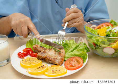 Asian senior woman patient eating pork chop stake and vegetable salad for healthy food in hospital. 128861803