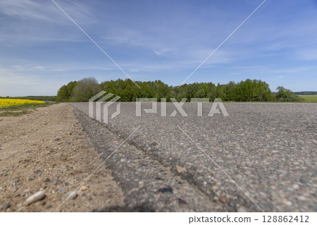 an asphalt road in a field on one side grows flowering rapeseed, and on the other side grows green grass against a blue sky with clouds 128862412