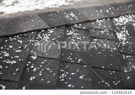 Close-up of black roof shingles covered with scattered hailstones during a hailstorm, showing texture and water droplets. Close-up of black roof shingles covered with scattered hailstones during a hailstorm, showing texture and water droplets. 128862810
