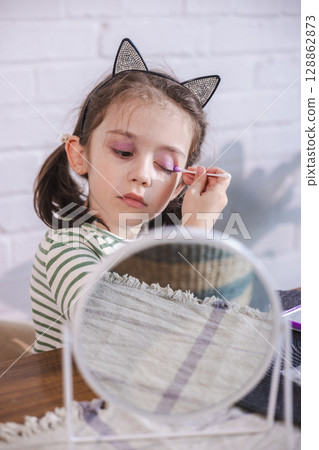 Young girl with cat ear headband applies bright pink eyeshadow with a cotton swab, deeply concentrated and enjoying her makeup play Young girl with cat ear headband applies bright pink eyeshadow with a cotton swab, deeply concentrated and enjoying her makeup play 128862873