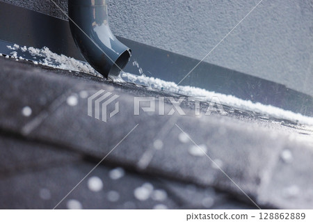 Water flowing from a gray rain gutter onto a dark roof covered with small hailstones during a storm. 128862889