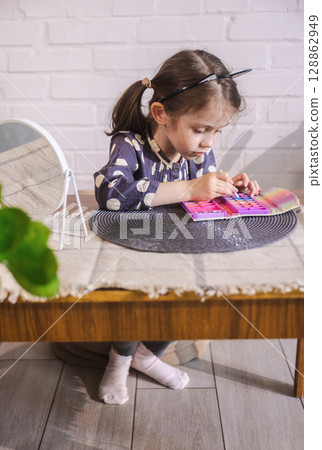 Little girl in playful cat-ear headband intently applying toy makeup at a table with a mirror 128862949