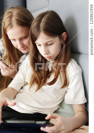 Two teenage girls lounging on a bed and playing separately, one on a tablet and the other on a smartphone, both completely absorbed in their screens Two teenage girls lounging on a bed and playing separately, one on a tablet and the other on a smartphone, both completely absorbed in their screens 128862956