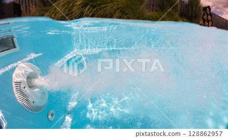 Underwater view of a jet stream creating intense bubbles in a backyard pool with decorative greenery and wooden fencing in the background. 128862957