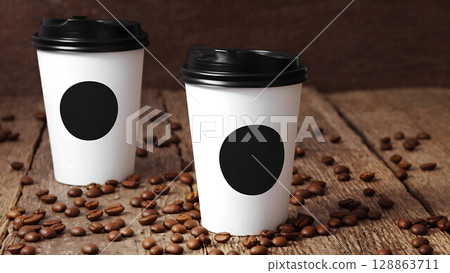 Two white coffee cups with black lids are placed on a wooden table, which is covered with coffee beans. The cups are filled with a white liquid, possibly coffee 128863711