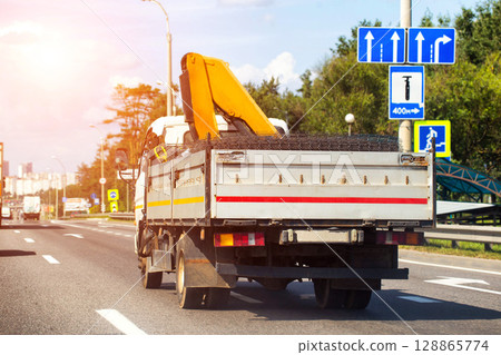 A dump truck with a hydraulic manipulator transports a metal mesh in the back along the road against the backdrop of the sun, cargo transportation 128865774