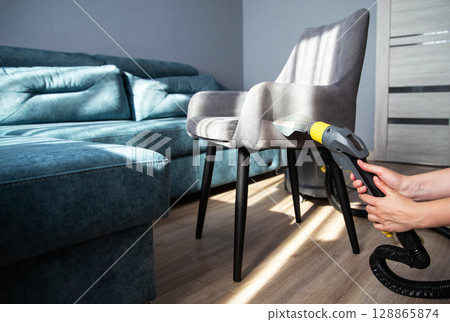 A girl s hand with a washing vacuum cleaner cleans the gray upholstery of a fabric chair in the apartment from dirt. Cleaning services, upholstered furniture cleaning 128865874