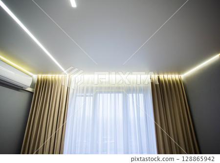 Beautiful designer stylish stretch ceiling with LED light lines against the background of a window with curtains in the living room. Option for lighting with light lines. 128865942