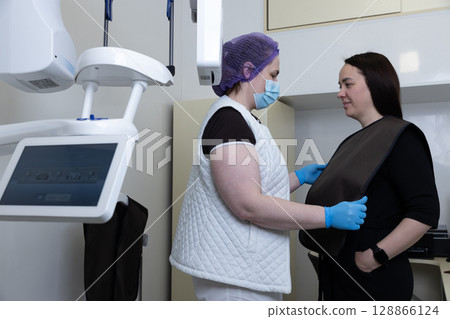 Patient Preparation for Medical Imaging Procedure: A Healthcare Professional Assists a Woman with a Protective Apron in a Modern Medical Setting 128866124
