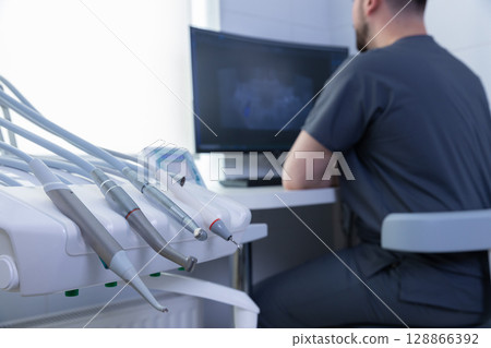 A Focused Dentist at Work in a Modern Dental Office with State-of-the-Art Equipment and a Computer Monitor Displaying Patient X-rays for Diagnosis and Treatment Planning 128866392