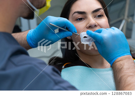A Dental Professional is Conducting an Oral Examination on a Patient Wearing a Protective Mask, Both Engaged in a Dental Procedure in a Modern Clinic Environment. A Dental Professional is Conducting an Oral Examination on a Patient Wearing a Protective Mask, Both Engaged in a Dental Procedure in a Modern Clinic Environment. 128867083