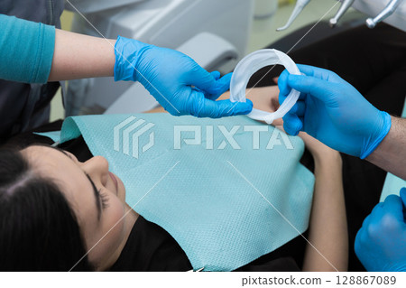 Patient in Dental Office Receiving Care, Dentists Prepare Dental Equipment and Accessories Including Protective Ring in Professional and Comfortable Setting 128867089