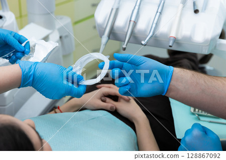 Dental professionals preparing for a procedure in a modern dental office, showcasing proper hygiene protocols with gloves and dental tools on the tray, ensuring patient comfort and safety. Dental professionals preparing for a procedure in a modern dental office, showcasing proper hygiene protocols with gloves and dental tools on the tray, ensuring patient comfort and safety. 128867092