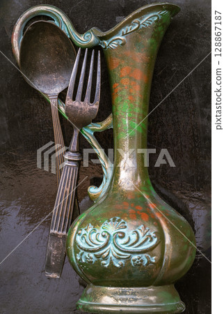 Black metal spoon and fork on Antique carved iron jug or vase with the Dark background. Black metal spoon and fork on Antique carved iron jug or vase with the Dark background. 128867187