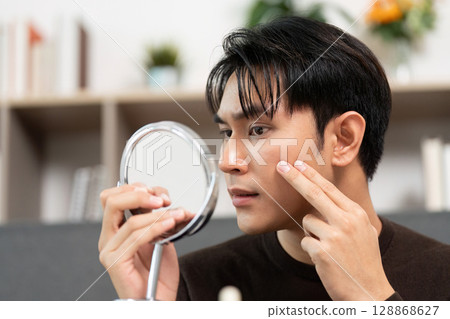 Skincare Routine. Young man examining skin with mirror for healthy complexion. 128868627