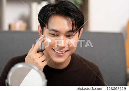 Skincare Routine. Young man applying moisturizer on face in front of mirror. 128868629