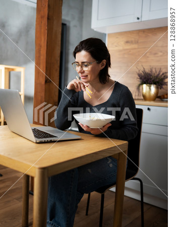 Happy Woman Eating Noodles And Reading A Message On Laptop At The Kitchen Table 128868957