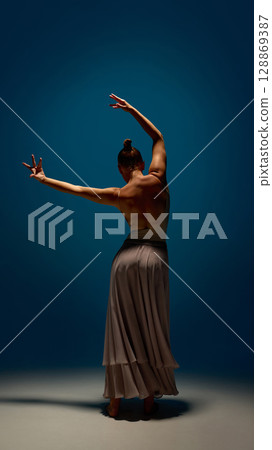 Infinite body. Female contemporary dancer standing in elegant pose with raised arms and long skirt under spotlight 128869387