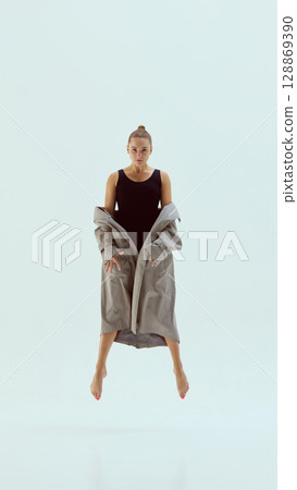 Contemporary dancer floating in mid-air with focused gaze and trench coat against light backdrop Contemporary dancer floating in mid-air with focused gaze and trench coat against light backdrop 128869390