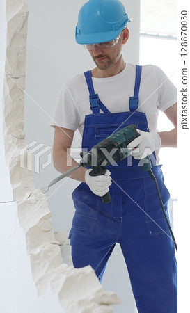 Man construction worker wearing blue overalls, hard hat and protective white gloves, is demolishing white wall with rotary hammer drill, generating dust, close up horizontal view. Renovation concept Man construction worker wearing blue overalls, hard hat and protective white gloves, is demolishing white wall with rotary hammer drill, generating dust, close up horizontal view. Renovation concept 128870030