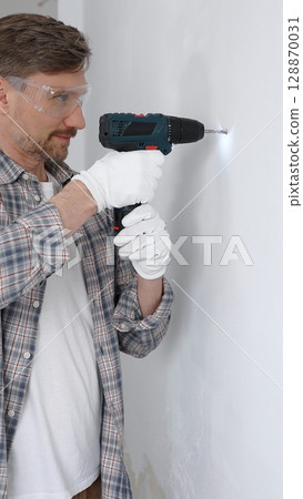 Middle aged man worker wearing grey checked shirt and white gloves, is drilling hole in a wall with cordless drill during home renovation work. Portrait view Middle aged man worker wearing grey checked shirt and white gloves, is drilling hole in a wall with cordless drill during home renovation work. Portrait view 128870031