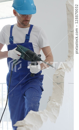 Male construction worker wearing blue overalls, protective helmet and white gloves, is demolishing white wall with rotary hammer drill, generating dust, close up horizontal view. Renovation concept 128870032