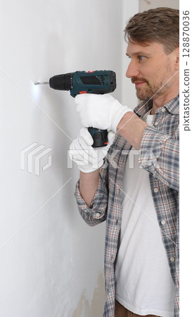 Middle aged male professional worker wearing grey checked shirt and white gloves, is drilling hole in a wall with cordless drill during home renovation work. Vertical portrait view 128870036