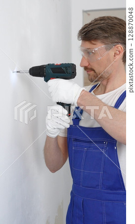 Professional male construction worker in a blue jumpsuit, safety goggles, and work gloves is screwing a screw into the wall using a cordless screwdriver, carrying out a careful home renovation 128870048