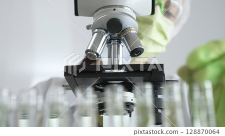 Scientist in green protective mask and gloves holding test tube with plant sample, researching botany and medical applications using a microscope. Medicine, healthcare and science concept Scientist in green protective mask and gloves holding test tube with plant sample, researching botany and medical applications using a microscope. Medicine, healthcare and science concept 128870064
