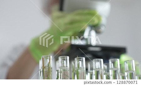Close up of test tubes with a scientist analyzing plant samples in test tubes with a microscope, conducting research in a laboratory. Medicine, healthcare and science concept 128870071