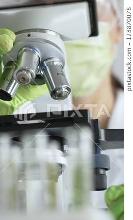 Close up of green gloved hands of a scientist adjusting a microscope in a laboratory, emphasizing the significance of scientific research and analysis. Medicine, healthcare and science concept 128870078
