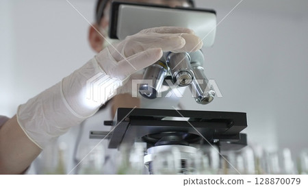 Female scientist wearing white protective gear analyzing test samples under microscope in contemporary research environment, close up of white gloved hand. Medicine and science concept Female scientist wearing white protective gear analyzing test samples under microscope in contemporary research environment, close up of white gloved hand. Medicine and science concept 128870079