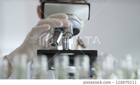 Female scientist wearing white protective gear analyzing test samples under microscope in contemporary research environment. Medicine, healthcare and science concept Female scientist wearing white protective gear analyzing test samples under microscope in contemporary research environment. Medicine, healthcare and science concept 128870111