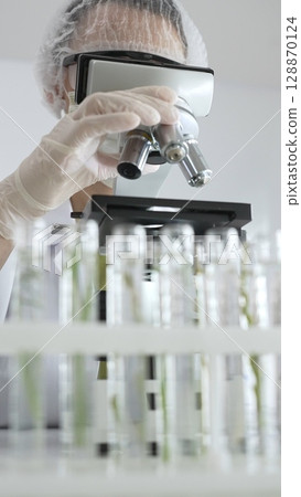 Scientist wearing green protective gear using microscope, analyzing test tubes in laboratory, conducting scientific research and development. Medicine, healthcare and science concept Scientist wearing green protective gear using microscope, analyzing test tubes in laboratory, conducting scientific research and development. Medicine, healthcare and science concept 128870124