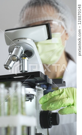 Close up of scientific precision involving green gloved hands adjusting microscope lens, blurred laboratory glassware highlighting medical investigation. Medicine, laboratory and science concept 128870148