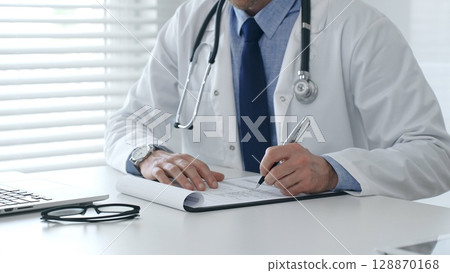 Close-up of male doctor's hands filling out medical form, sitting at desk with laptop and glasses, wearing white coat and stethoscope in medical office. Medicine and healthcare concept 128870168