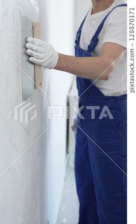 Man construction worker wearing protective gloves and blue construction coveralls, spreading plaster smoothly across wall using professional drywall taping knife 128870171