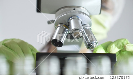 Close up of green gloved hands of a scientist adjusting a microscope in a laboratory, emphasizing the significance of scientific research and analysis. Medicine, healthcare and science concept 128870175