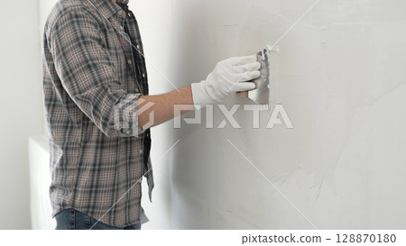 Unknown construction worker man wearing gloves and plaid shirt is using a putty knife to apply plaster on a white wall, during a home renovation or construction project Unknown construction worker man wearing gloves and plaid shirt is using a putty knife to apply plaster on a white wall, during a home renovation or construction project 128870180