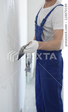 Unknown man construction worker wearing gloves and blue overalls is using a putty knife to apply plaster on a white wall, performing home renovation and improvement Unknown man construction worker wearing gloves and blue overalls is using a putty knife to apply plaster on a white wall, performing home renovation and improvement 128870194