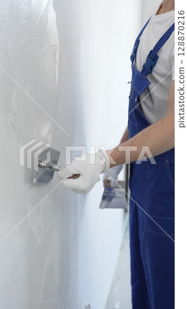 Professional construction worker wearing blue work overall is using a finishing trowel to carefully apply plaster on a wall, demonstrating expertise in home renovation Professional construction worker wearing blue work overall is using a finishing trowel to carefully apply plaster on a wall, demonstrating expertise in home renovation 128870216