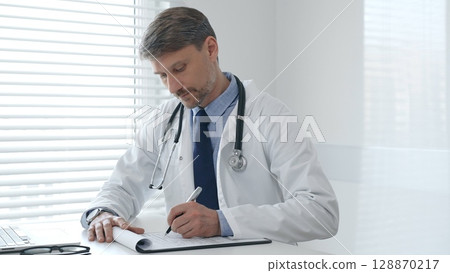 Male physician wearing a lab coat and stethoscope, sitting at a desk in a medical office, focused on using a laptop while taking notes on a clipboard for patient care and treatment Male physician wearing a lab coat and stethoscope, sitting at a desk in a medical office, focused on using a laptop while taking notes on a clipboard for patient care and treatment 128870217