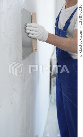 Man construction worker wearing protective gloves and blue construction coveralls, spreading plaster smoothly across wall using professional drywall taping knife Man construction worker wearing protective gloves and blue construction coveralls, spreading plaster smoothly across wall using professional drywall taping knife 128870230