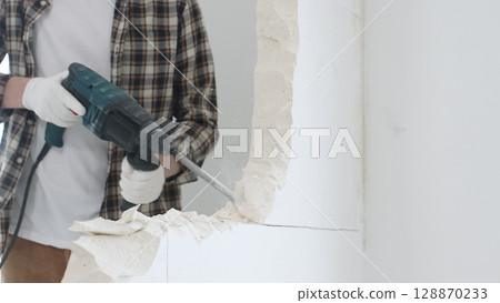 Close up of unrecognizable male construction worker wearing beige checkered shirt and protective gloves, is demolishing white wall with rotary hammer drill, generating dust, close up view. Renovation Close up of unrecognizable male construction worker wearing beige checkered shirt and protective gloves, is demolishing white wall with rotary hammer drill, generating dust, close up view. Renovation 128870233