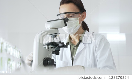 Female scientist wearing a lab coat, face mask, and safety glasses carefully adjusts a microscope, conducting research in a brightly lit laboratory setting. Medicine, healthcare and science concept Female scientist wearing a lab coat, face mask, and safety glasses carefully adjusts a microscope, conducting research in a brightly lit laboratory setting. Medicine, healthcare and science concept 128870234
