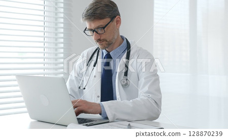 Focused mature male doctor wearing eyeglasses and lab coat with stethoscope is typing on laptop keyboard while sitting at desk in bright medical office Focused mature male doctor wearing eyeglasses and lab coat with stethoscope is typing on laptop keyboard while sitting at desk in bright medical office 128870239